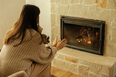 Kır odasında şöminede elleri ısıtan rahat kazaklı bir kadın. Çiftlik evindeki şöminede oturan şık bir kadın. Kışın şömine ısıtması, gaz ve elektriğe alternatif.