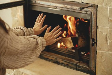Kışın odun sobasıyla ısıtma evi. Çiftlik evindeki kır evinde yanan şöminede elleri ısıtan bir kadın. Gaz ve elektriğe alternatif olarak şömine ısıtma