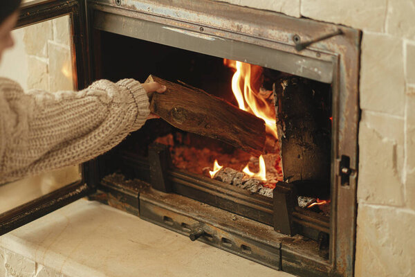 Heating house in winter with wood burning stove. Woman throwing firewood into burning fireplace in rustic room in farmhouse. Fireplace heating alternative to gas and electricity