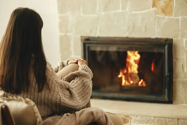穿着舒适毛衣的时髦女人在乡村房间壁炉边的椅子上热身 冬天用烧木柴的炉灶取暖 年轻的女性在农舍的壁炉边坐着休息 — 图库照片