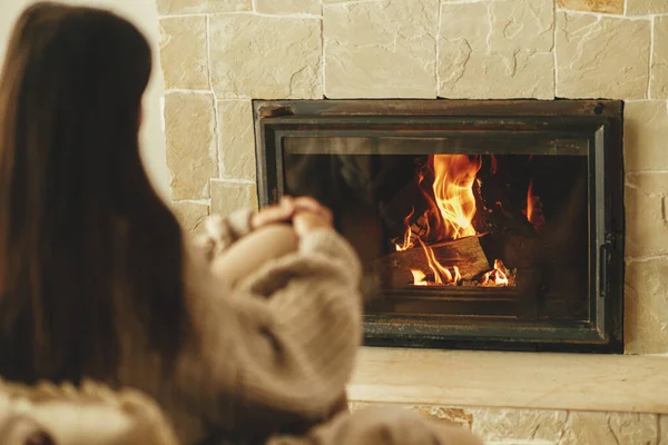 冬天用烧木柴的炉灶取暖 穿着舒适毛衣的时髦女人在乡村房间壁炉边的椅子上热身 年轻的女性在农舍的壁炉边坐着休息 — 图库照片