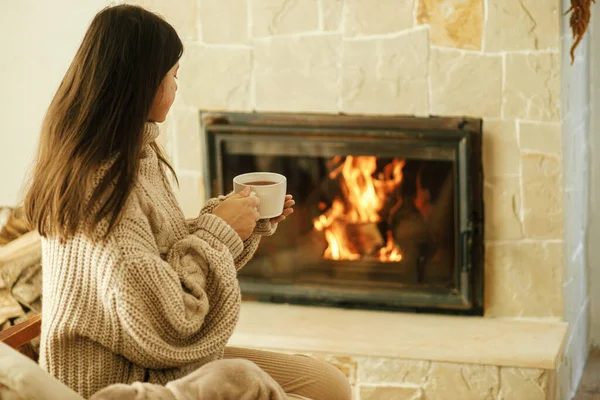 穿着舒适毛衣的女人在乡村房间的壁炉边端着一杯热茶 冬天用烧木柴的炉灶取暖 年轻时尚的女性在农舍的壁炉前休息 — 图库照片