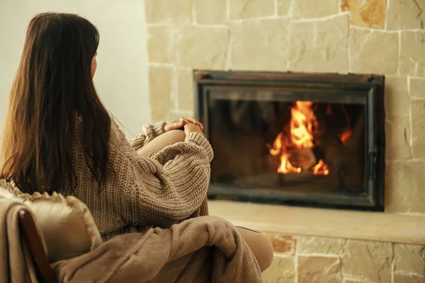 穿着舒适毛衣的时髦女人在乡村房间壁炉边的椅子上热身 冬天用烧木柴的炉灶取暖 年轻的女性在农舍的壁炉边坐着休息 — 图库照片