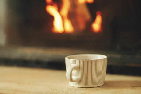 Stylish cup of warm tea on background of burning fireplace close up, autumn winter hygge. Heating house with wood burning stove. Relaxing and warming up at cozy rustic fireplace