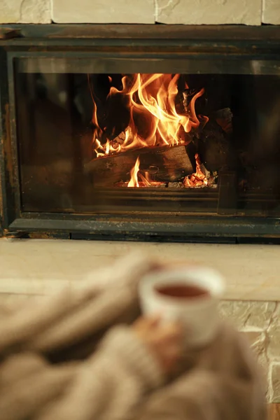 Heating house with wood burning stove. Burning fireplace close up and blurred hands with cup of warm tea , autumn hygge. Relaxing and warming up at cozy rustic fireplace