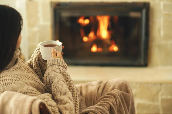 穿着舒适毛衣的女人在乡村房间的壁炉边端着一杯热茶 冬天用烧木柴的炉灶取暖 年轻时尚的女性在农舍的壁炉前休息 — 图库照片