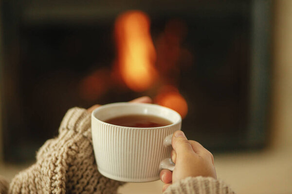 Hands in cozy sweater holding cup of warm tea on background of burning fireplace close up, autumn hygge. Heating house with wood burning stove. Relaxing and warming up at rustic fireplace