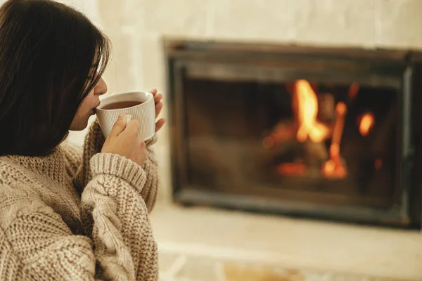穿着舒适毛衣的女人在乡村房间的壁炉边喝杯热茶 冬天用烧木柴的炉灶取暖 年轻时尚的女性在农舍的壁炉前休息 — 图库照片
