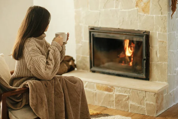 暖炉で暖かいお茶のカップを保持居心地の良いセーターの女性は 秋の巨大 薪ストーブで家を暖める 若いスタイリッシュな女性は素朴な農家の暖炉で居心地の良い椅子でリラックス — ストック写真
