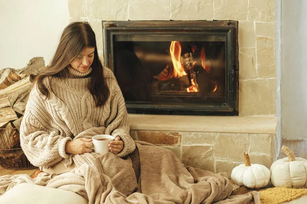 女人带着一杯热茶在壁炉边放松 秋天的乡下人 用烧木柴的火炉加热房子 乡村农舍壁炉旁穿着舒适毛衣的年轻时尚女性 — 图库照片