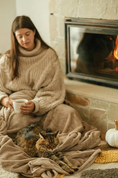 Cute cat and woman with cup of tea sitting together on cozy blanket at fireplace. Adorable tabby kitty relaxing together with owner at burning fireplace in rustic farmhouse