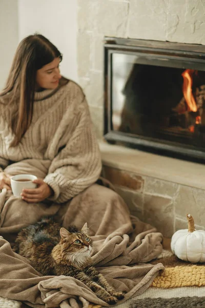 Cute cat and woman with cup of tea sitting together on cozy blanket at fireplace. Adorable tabby kitty relaxing together with owner at burning fireplace in rustic farmhouse