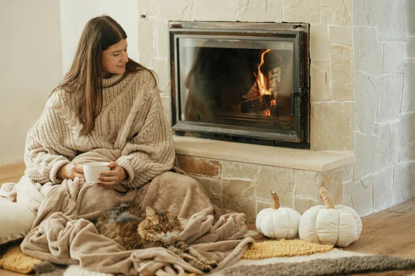 Woman with cup of tea and cute cat sitting together on cozy blanket at fireplace. Adorable tabby kitty relaxing together with owner at burning fireplace in rustic farmhouse