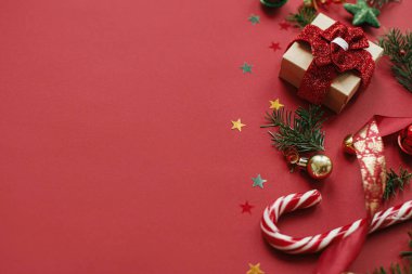 Modern Noel kompozisyonu. Şık bir Noel hediyesi, takılar, köknar dalları, kırmızı arka planda konfeti. Mevsimlik tebrik kartı, mesaj için yer. Mutlu Noeller! Kış tatili afişi