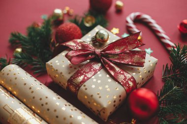 Mutlu Noeller ve mutlu tatiller! Modern Noel kompozisyonu. Şık bir Noel hediyesi, ambalaj kağıdı, köknar dalları, baston şekerler ve kırmızı arka planda mücevherler. Bayram bayrağı