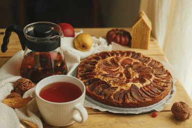 Atmosferik sonbahar natürmort. Tarçınlı, mumlu, sonbahar yapraklı, balkabaklı ve fındıklı taze elmalı turta ile sade ahşap masada, şık bir fincanda ılık çay. Hygge evde, rahat bir an.