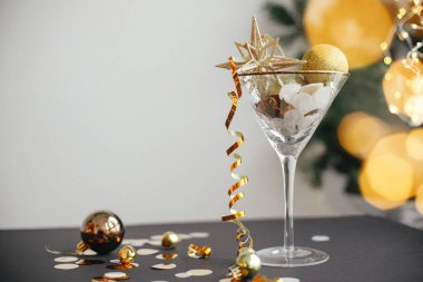 New year party creative concept. Stylish christmas glitter star in champagne glass, golden confetti and baubles on background of festive lights. Merry Christmas and Happy New Year!