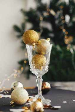 Merry Christmas and Happy New Year! Stylish christmas glitter baubles in champagne glass and golden decorations on dark table. Creative new year party composition. Moody holiday banner