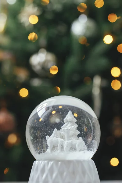 Stylish christmas snow globe on background of christmas tree in lights in festive decorated boho room. Snowy white snow globe against lights bokeh. Merry Christmas! Atmospheric winter time