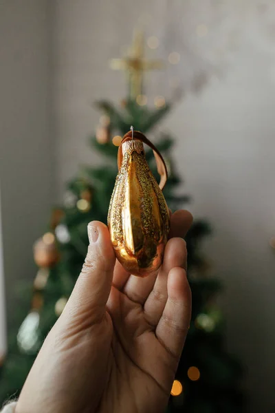 Christmas vintage ornament in hand on background of christmas tree in lights in festive decorated boho room. Person holding beautiful vintage bauble. Merry Christmas! Atmospheric winter time