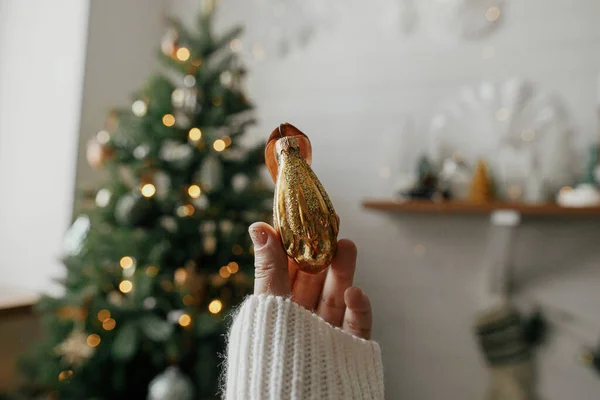 Christmas vintage ornament in hand on background of christmas tree in lights in festive decorated boho room. Person holding beautiful vintage bauble. Merry Christmas! Atmospheric winter time