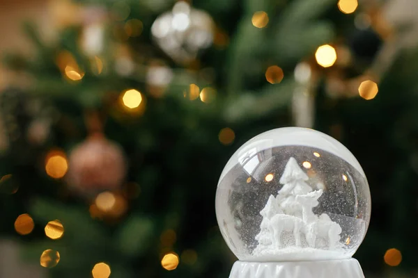 Stylish christmas snow globe on background of christmas tree in lights in festive decorated boho room. Snowy white snow globe against lights bokeh. Merry Christmas! Atmospheric winter time