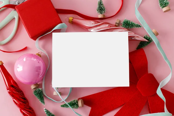 Christmas card mockup, pink flat lay. Empty card with space for text and red bow, little trees, baubles, ribbons on pink background. Modern creative template, seasons greetings