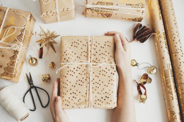 Merry Christmas! Hands holding stylish golden christmas gifts flat lay. Person preparing modern christmas gift boxes, festive paper, golden ornaments and thread composition on white wood
