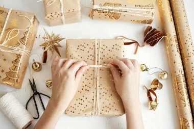 Mutlu Noeller! Şık altın noel hediyelerini saran eller dümdüz uzanıyordu. Modern noel hediye kutuları hazırlayan kişi, bayram kağıdı, altın süsler ve beyaz ahşap iplik kompozisyonu