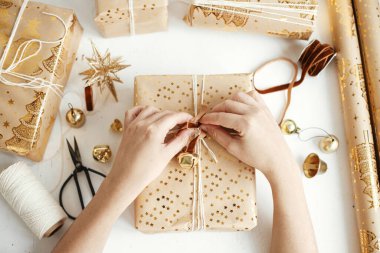 Mutlu Noeller! Şık altın noel hediyelerini saran eller dümdüz uzanıyordu. Modern noel hediye kutuları hazırlayan kişi, bayram kağıdı, altın süsler ve beyaz ahşap iplik kompozisyonu