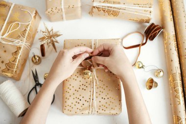 Şık altın noel hediyelerini saran eller dümdüz uzanıyordu. Modern noel hediye kutuları hazırlayan kişi, bayram kağıdı, altın süsler ve beyaz ahşap üzerine iplik kompozisyonu. Mutlu Noeller.!