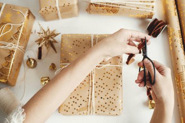 Şık altın noel hediyelerini saran eller dümdüz uzanıyordu. Modern noel hediye kutuları hazırlayan kişi, bayram kağıdı, altın süsler ve beyaz ahşap üzerine iplik kompozisyonu. Mutlu Noeller.!