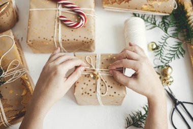 Şık Noel hediyelerini paketleyen eller dümdüz uzanıyordu. Modern noel hediye kutuları hazırlayan kişi, bayram kağıdı, altın süsler ve beyaz ahşap üzerine iplik kompozisyonu. Atmosfer zamanı, Mutlu Tatiller!