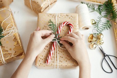 İyi tatiller! Şık Noel hediyesi paketleri düzleştiren eller. Modern Noel hediye kutusu hazırlayan kişi şeker kamışı, bayram kağıdı, beyaz ahşap üzerine altın süsler.
