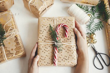 İyi tatiller! Şık Noel hediyesi paketleri düzleştiren eller. Modern Noel hediye kutusu hazırlayan kişi şeker kamışı, bayram kağıdı, beyaz ahşap üzerine altın süsler.