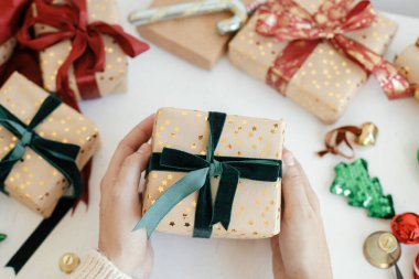 Hands wrapping stylish christmas gift. Person preparing modern gift box with green ribbon, golden wrapping paper, ornaments on white background. Merry Christmas! Atmospheric winter time