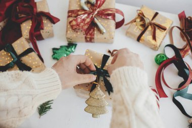 Mutlu Noeller! Eller şık bir Noel hediyesi hazırlıyor. Modern hediye kutusunu hazırlayan kişi altın ağaç, yeşil kurdele, ambalaj kağıdı, beyaz arka planda süsler. Atmosferik kış zamanı