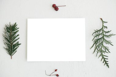 Christmas card mock up. Empty greeting card flat lay with fir branches, berries and christmas decorations on rustic white table. Postcard template with space for text. Happy Holidays!