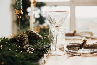 Şık bir Noel sofrası. Tabağında zil olan keten peçeteler, klasik çatal bıçak takımı, şarap kadehi, altın ışıklı köknar dalları, çam kozalakları ve masada mum. Atmosferik Tatil Kahvaltısı