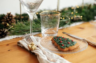 Noel sofrası. Tabakta Noel ağacı kurabiyesi, çanlı keten peçete, klasik çatal bıçak takımı, bardaklar, altın ışıklı köknar dalları ve masa üzerinde çam kozalakları. Modern Tatil Düzenlemesi