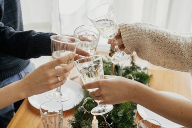 Noel ziyafeti. Şarap kadehli eller arka planda altın ışıklı ve mumlu köknar dalları olan şık masa ortamında kadeh kaldırıyor ve tıklıyor. Arkadaşlar şampanyayla kutlama yapıyor