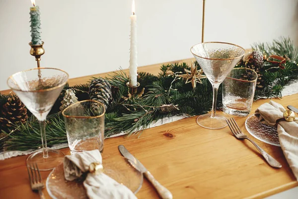 スタイリッシュなクリスマステーブルの設定 皿の上に鐘を持つリネンナプキン ヴィンテージカトラリー ワイングラス 黄金のライトとモミの枝 テーブルの上に松のコーンとキャンドル 大気の休日ブランチ — ストック写真
