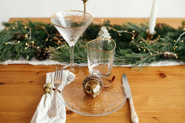 Stylish Christmas table setting. Linen napkin with bell, acorn on plate, vintage cutlery, wineglass, fir branches with golden lights, pine cones and candle on table.  Holiday brunch