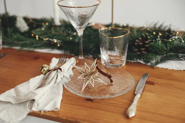Stylish Christmas table setting. Linen napkin with bell, star on plate, vintage cutlery, wineglass, fir branches with golden lights, pine cones and candle on table.  Holiday brunch