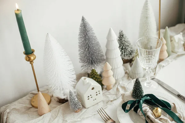 stock image Christmas table setting. Stylish napkin with ribbon and bell on plate, vintage cutlery, modern festive christmas trees and houses on white rustic table. Holiday arrangement of table