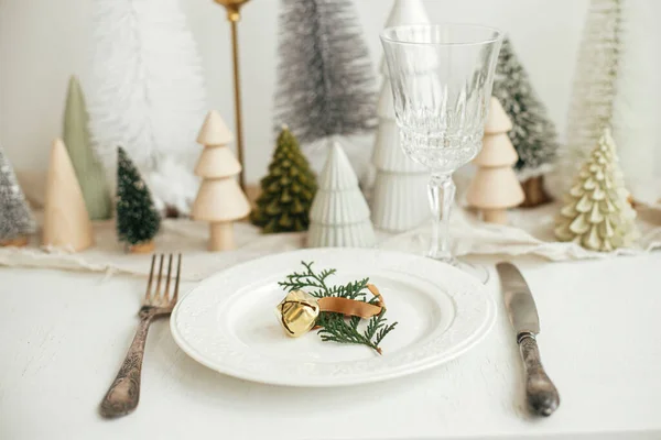 スタイリッシュなクリスマステーブルの設定 板の上に鐘を持つ杉の枝 ヴィンテージのカトラリー ワイングラス モダンなクリスマスツリーや白素朴なテーブルの上の家 休日のブランチ 新年のお祝い — ストック写真