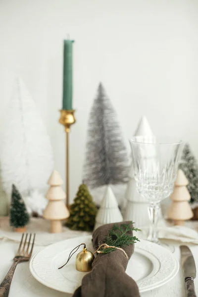 スタイリッシュなクリスマステーブルの設定 プレート上の鐘とモミの綿のナプキン ヴィンテージカトラリー ワイングラス モダンなクリスマスツリーと白い素朴なテーブルの上にキャンドル 休日ブランチ — ストック写真