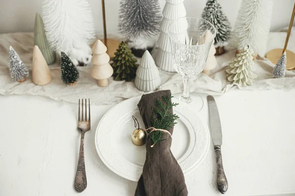 クリスマステーブルのセッティング プレート上の鐘やモミとスタイリッシュなナプキン ヴィンテージカトラリー 白い素朴なテーブルの上にお祝いの小さなクリスマスツリー テーブルの休日の配置 トップビュー — ストック写真