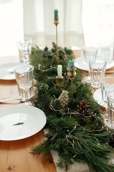 Stylish Christmas table setting. Rosemary branch on plate, vintage cutlery, wine glass, candles, fir branches with golden lights, pine cones on table. Holiday dinner, atmospheric table set