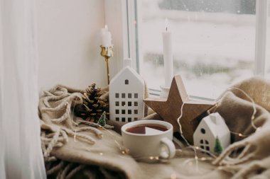 Karlı bir günde sıcak bir ev. Sıcak bir fincan çay, mumlar, ışıklar, küçük noel evleri ve ağaçlar, pencere kenarındaki battaniyenin üzerindeki ahşap yıldız. Kış kızarıklığı. Atmosferik İskandinav havası
